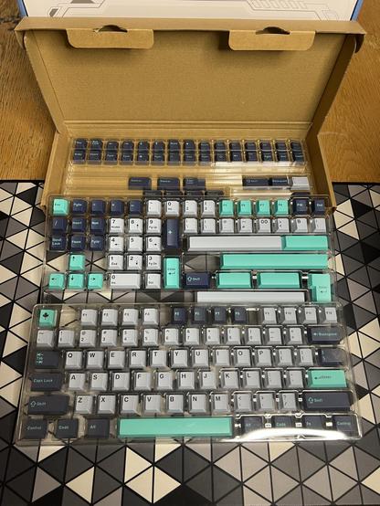 An open box of keycaps showing grey, teal and dark grey cherry style keycaps laid out in plastic trays.