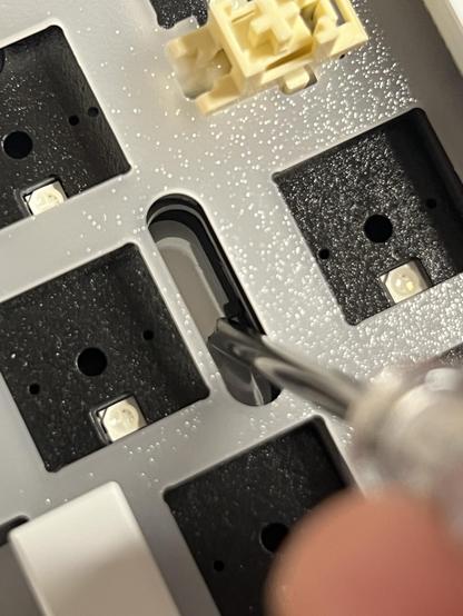 A screwdriver poking through the plate and PCB of a mechanical keyboard to reach a little switch on the reverse side.