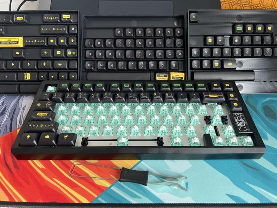A keyboard with unpopulated switches sat in front of three trays of assorted black keycaps with green, yellow and white text.