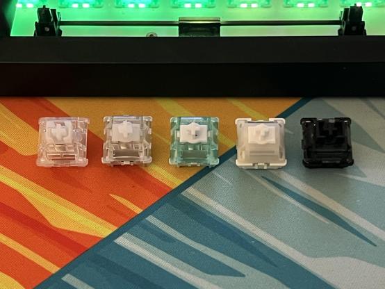 Four mechanical keyboard switches arranged in a line in front of a mechanical keyboard with an unpopulated spacebar.