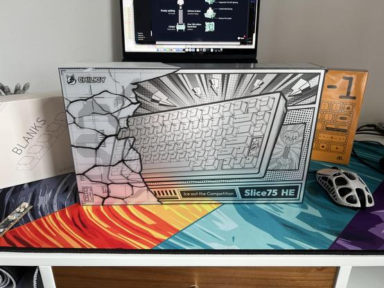 A shiny silver mechanical keyboard box stood upright on a desk. Two boxes of keycaps peek out from behind it.