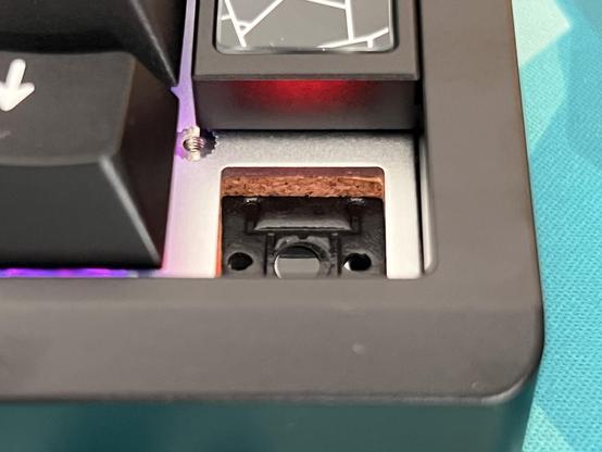 The corner of a keyboard with a keycap and switch removed, showing a silver aluminium plate, a cork layer and the PCB beneath.