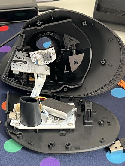A trackball mouse unscrewed and open upon a desk. The top and bottom shells are joined by two ribbon cables that attach the mouse wheel and sensor PCBs. A comically small LiPo battery sits on the bottom PCB at a jaunty angle.