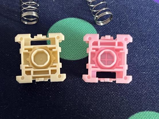 Two switch bottom housings. One a sort of beige and one pink. The pink one - the “Deep Clacky” - has a viable cross shape in the bottom of its stem housing and plastic so thin that the deskmat colour slightly shows through.