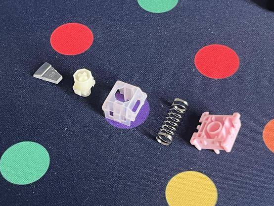 Another disassembled mechanical keyboard switch, its parts arrayed out on a dotty deskmat. They comprise a little light pipe, the stem, the top housing, a spring and the bottom housing.