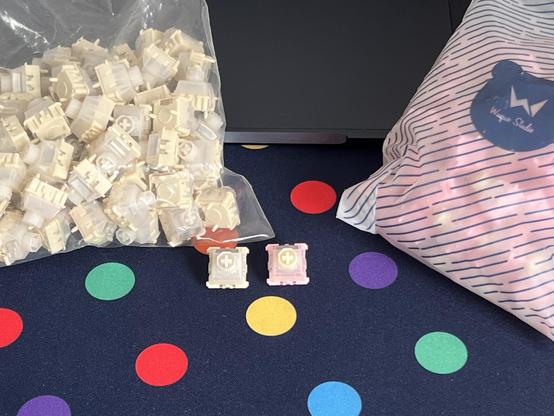 Two bags of keyboard switches sat atop of a desk. A switch from each is placed in front.