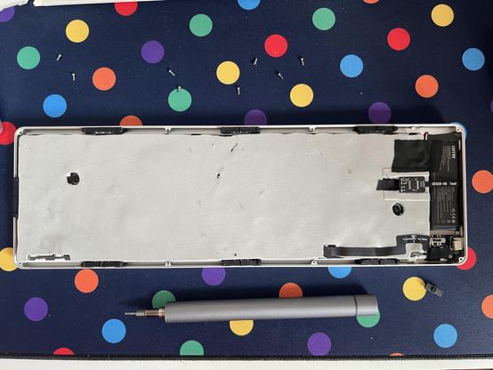 A low profile compact keyboard with the back unscrewed revealing a set of black flat flex cables and a small black battery on one end, and an expanse of light grey foam damping over the rest.