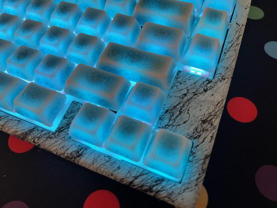 A closeup of the top of a mechanical keyboard, with blue crazed ceramic keycaps illuminated blue. The top shell has been replaced with a blue painted 3D printed layer, decorated with marble effect spray paint. It… mostly works.