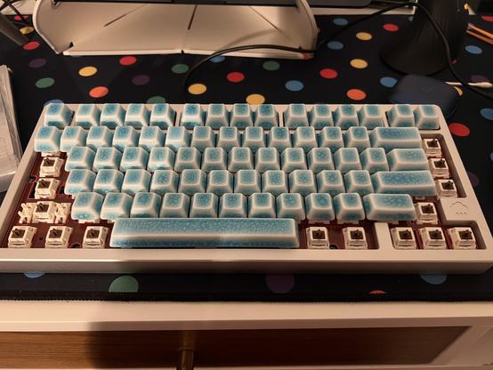 A chunky, 75% aluminium keyboard being populated with blue and white crazed ceramic keycaps. Unpopulated caps reveal a red FR4 plate. The blocker on the right-hand edge of the keyboard has a little rain cloud logo.