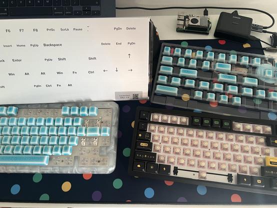 The back of a keycap box with labels describing which keys in the included tray are which. Beneath it are two keyboards with the ceramic caps populated on the left most one.