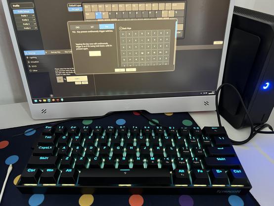 A black 60% mechanical keyboard with backlit letters sitting in front of a small tabletop monitor showing the advanced key editor in the software.