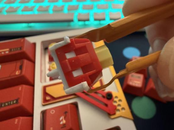 A hand holding a small orange metal switch puller which is grasping a white, red and yellow keyboard switch. A matching white, red and yellow keyboard is blurred the background.