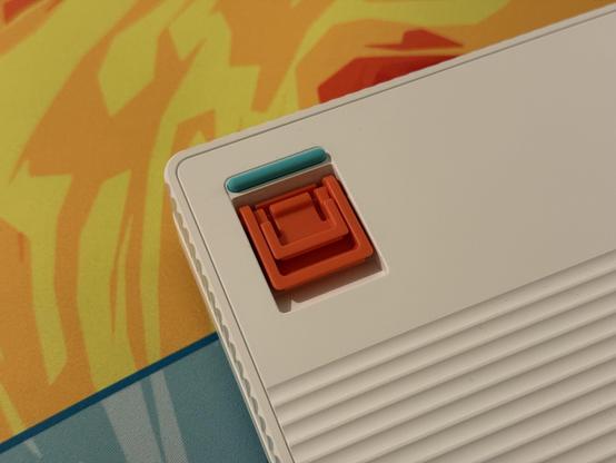 The bottom of a white mechanical keyboard showing a bright red, two-stage flip out foot for tilt adjustment, next to an accent teal rubber foot.