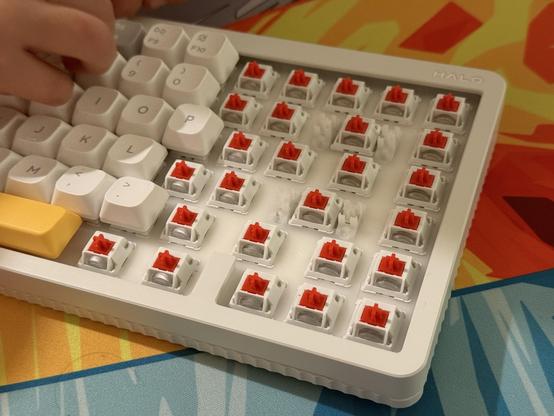 A white mechanical keyboard with several keycaps removed and a tiny hand removing more. The switches are exposed, white with red stems.