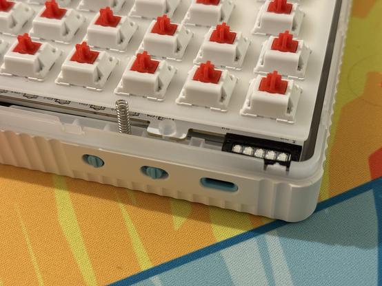 The bottom half of a white mechanical keyboard with the metal top shell removed. Some LEDs and a sticky-up metal spring are revealed.