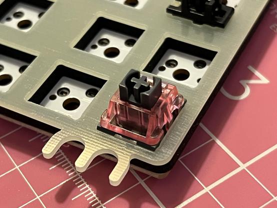 A single translucent keyboard switch plugged into the corner of a plate, foam and PCB assembly, holding all three together.