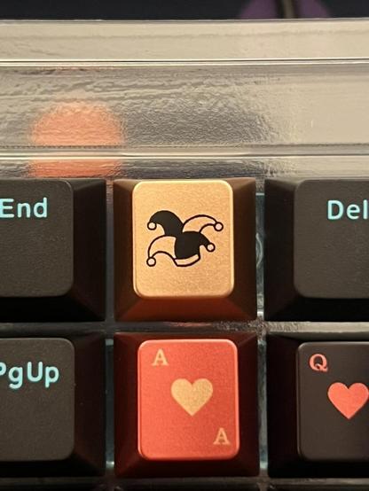 A closeup of six keycaps from two different sets. The top middle is a very distinct Jester hat design in black on a gold keycap. It's unmistakably similar to one from a child kit (novelties) of an expensive, limited run keycap set.
