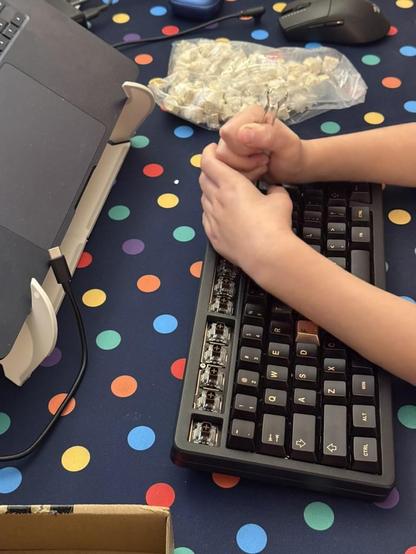 A black mechanical keyboard. Obscuring it are small hands holding a keycap remover and pulling up a key. A bag of switches lies ot one side.