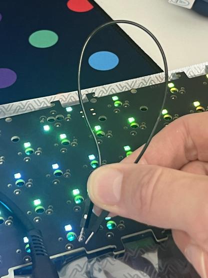 A keyboard lit up with blue green LEDs and a long black curved wire - for connecting the two sides of a hotswap switch socket - held in front.