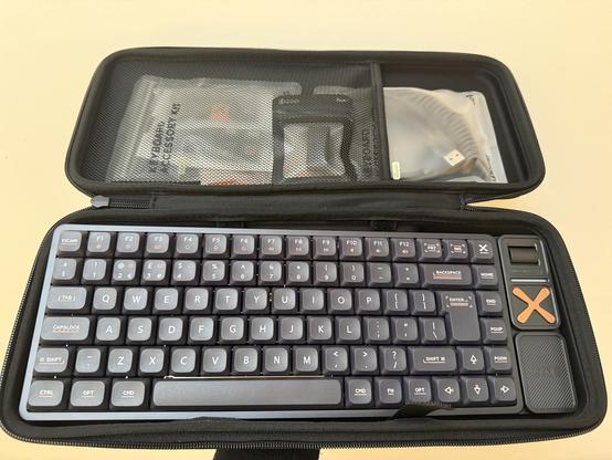 An IQUNIX 75% mechanical keyboard in a soft case with keycaps, coiled cable and other accessories in the top half. The keyboard is low profile with a copper cross-shaped four way media nav and a roller volume control in a self-contained little detail on the right.