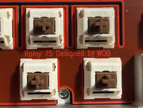 A cherry red keyboard plate with bright red edge outlines. Text in between some switches reads “Rainy 75 Designed by WOB.”