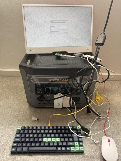 A mess of wires plugged into a portable battery. On top a Pi Zero is running and connected to a monitor. In front is a mechanical keyboard with cool green hacker style keycaps and a very clashing red and white Pi mouse.