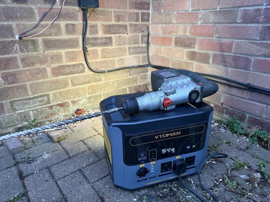 A huge SDS drill sat on top of a portable battery. The battery is casting light on the wall behind it, illuminating the work area in the waning late afternoon sun.