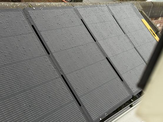 An awkward angle photo showing four large solar panels connected together with folding joints and arrayed over a pitched roof. They are spotted with rain.