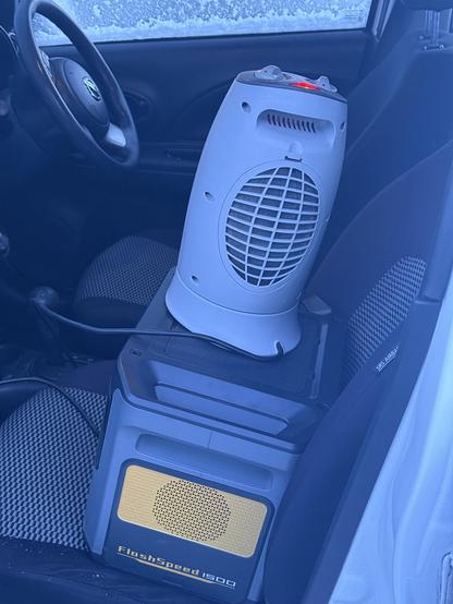 A small tower fan heater sat on top of a large portable battery. Both are perched on the passenger seat of a car which, from the snow on the window in the background and the blue cast over everything, is clearly icy and cold.