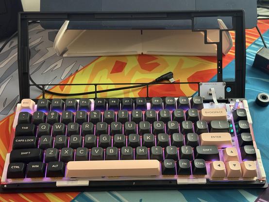 The top shell of a mechanical keyboard lifted off and propped up to one side. A short LCD ribbon cable joins it to the bottom.