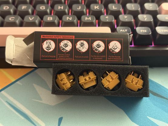 Four yellow mechanical keyboard switches nestled in a foam insert. The cardboard box from whence it came is sat next to it and features instructions for removing and replacing switches.