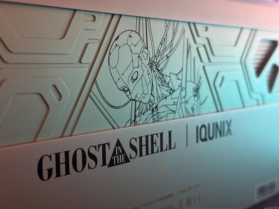The bottom of the IQUNIX Shell Core EV63, with a line drawing of a cyborg suspended by straps and connected to all manner of wires. Detailed thematically relevant angular patterns are milled into the metal which is a delicate hue of green.