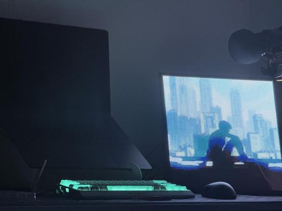 The IQUNIX Shell Core EV63 lit up in green. Behind it is a sleeping laptop shrouded in shadow. To the right is a bright display of some sort with a screenshot from the 1996 movie depicting the Major silhouetted against a cityscape.